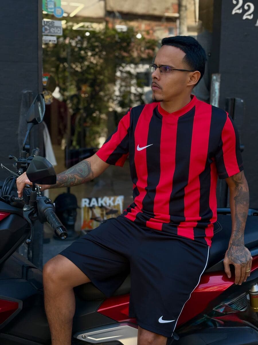 Cria de óculos e camisa listrada preta e vermelha, na moto na frente da loja